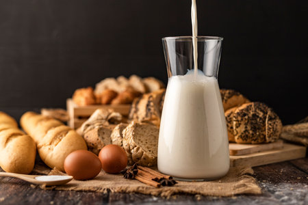 Pour the milk in a glass bottle placed on the sack. A lot of bread and eggs lay on a wooden table.