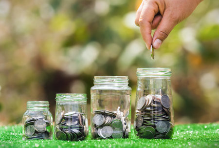 hand holding coins putting into jug glass with money stack on green grass. concept saving finance and accounting conceptの写真素材