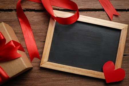 Selective focus of gift box with red ribbon, chalkboard and love shaped paper over rustic wooden table, top view flat lay.の写真素材