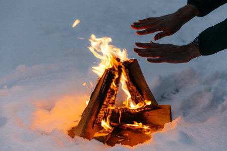 Unrecognizable male warms hands on fire in forest during cold winter, tries to warm himself, being cold spend time on cold snow and frosty weather. Burning flame on ground covered with snow.の写真素材