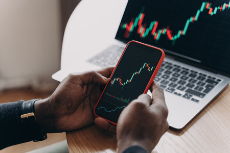 Cropped photo of male hands holding smartphone in front of computer screen analyzing financial stock...