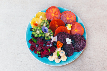 Top view of tropical fruit assortment on plate against white background. Fresh slices of dragon fruit, pitaya, kiwi, banana, blueberry decorated with small pansy. Bright colored healthy fruitの写真素材