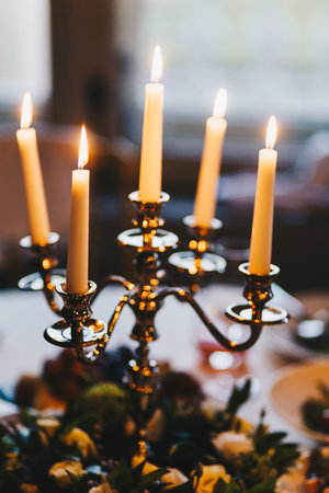 Shot of antique candlestick with five burning candles stand on table against light blurred background with floral arrangement. Lit candles in dining roomの写真素材