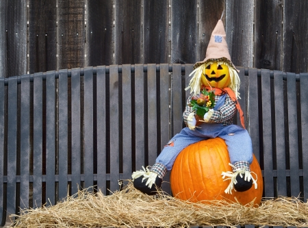 Scarecrow sitting on a pumpkin and hay. Gray iron bench and wooden fence on the background.の写真素材