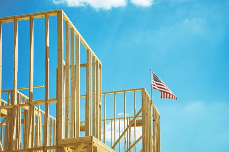American flag waving in the wind on the top of a construction home framing. Blue sky and white...