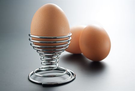 	Egg placed in a metal spiral egg-holder arranged with two boiled eggs on gray gradient backgroundの写真素材