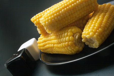 Freshly cooked corn cobs close-up arranged in a black ceramic plate with salt and pepper shakers on dark background の写真素材