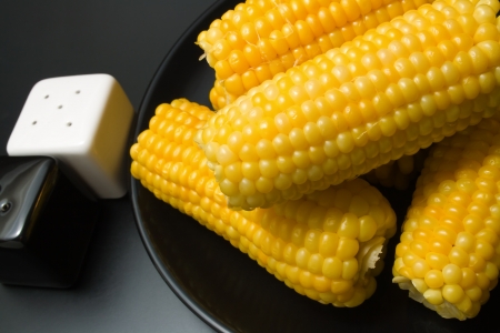 	Corn cobs. Freshly cooked corn cobs close-up arranged in a black ceramic plate with salt and pepper shakers on dark backgroundの写真素材