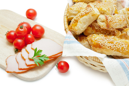 Freshly baked puff pastry patties sprinkled with sesame seeds in a wicker basket closeup arranged with cherry tomatoes ham and parsley  on white background.の写真素材