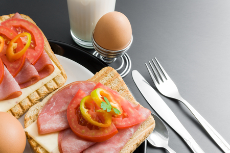 Breakfast. Pork tenderloin sandwiches, cheese, tomato, pepper and parsley  in ceramic plates arranged with boiled eggs placed in a metal stand, spoon, fork, knife and glass of milk close up on gray neutral gradient background.の写真素材