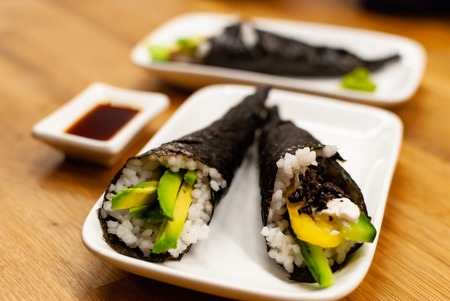 Side view Side view of homemade vegan Temaki-Sushi filled with avocado on a small plate on a wooden table with soy sauce and wasabiの写真素材