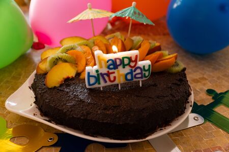 A home made vegan chocolate cake with fresh fruit, small umbrellas, balloons and a candle saying "Happy Birthday"の写真素材