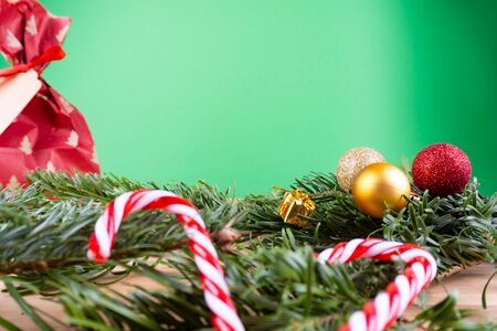 Green fir branches with Christmas ornaments (Christmas baubles, candy canes, presents, fir cone) on a wooden table in front of a green background with copy spaceの写真素材