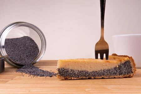 A piece of vegan cheesecake with poppy seeds together with a fork, a cup of coffee and poppy seeds on a wooden table - side viewの写真素材