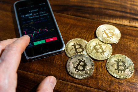 Smartphone with Bitcoin trading chart on the screen. Holding in hand a gold Bitcoin Cash coin. Trading on the cryptocurrency exchange.の写真素材