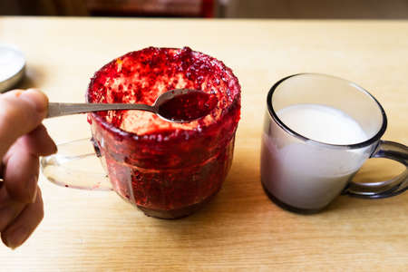 glass jar with raspberry jam. cup of milk or yogurt on wooden table. holding a spoon with raspberry jam.の写真素材