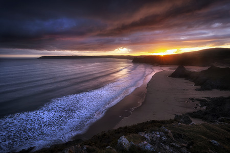 Sun setting behind the famous rock face of Three Cliffs Bay on the Gower peninsula in Swansea, South...