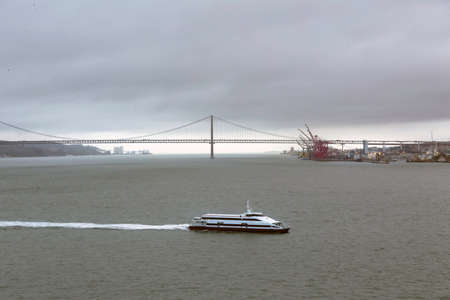Editorial Lisbon, Portugal - March 21, 2022: A ferryboat crossing the Tagus river near the Ponte 25 de Abril suspension bridge.のeditorial素材