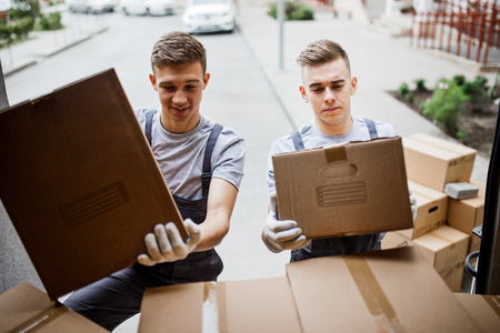 Two young handsome smiling movers wearing uniforms are unloading the van full of boxes. House move, mover service.の写真素材
