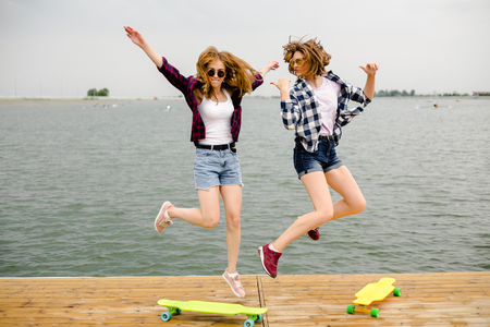 Two cheerful happy skater girls in hipster outfit having fun on a wooden pier during summer vacationの写真素材