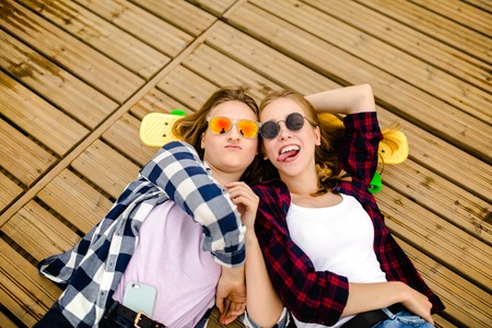 Two stylish young urban girls with longboards lie on the wooden flooring in the street. Friends have fun and spend time together in the summer during the holidaysの写真素材