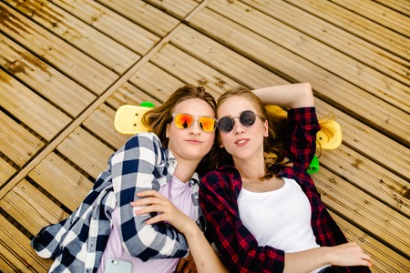 Two stylish young urban girls with longboards lie on the wooden flooring in the street. Friends have fun and spend time together in the summer during the holidaysの写真素材