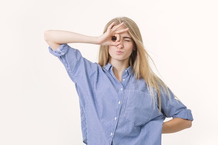 Portrait of cute joyful blonde girl, looking at the camera through fingers in okay gesture