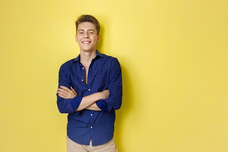 Friendly looking cheerful european guy wearing blue shirt smiling standing over yellow background with crossed handsの写真素材