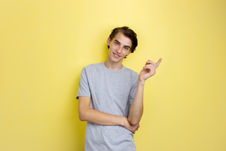Cheerful handsome young thin dark-haired guy with blue eyes wearing gray t-shirt pointing left,...