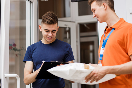 A person wearing an orange T-shirt is delivering a parcel to a satisfied client, who is putting his signature on the receipt. Friendly worker, high quality delivery service.の写真素材