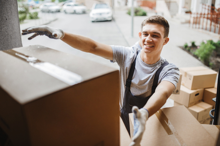 A young handsome smiling mover wearing uniform is reaching for the box while unloading the van full of boxes. House move, mover service.の写真素材