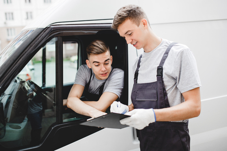 A young handsome guy wearing uniform is pointing at the clipboard while sitting in the car. Another worker wearing uniform is looking at the clipboard. House move, mover service.の写真素材