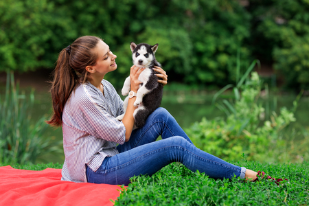 A beautiful smiling woman with a ponytail and wearing a striped shirt is cuddling with  a sweet husky puppy while resting on the red blanket on the lawn. Love and care for pets.の写真素材