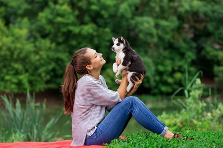 A beautiful smiling woman with a ponytail and wearing a striped shirt is cuddling with  a sweet husky puppy while resting on the red blanket on the lawn. Love and care for pets.の写真素材