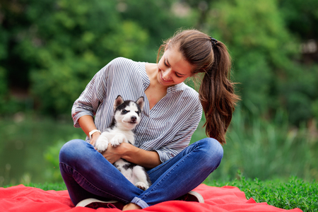 A beautiful smiling woman with a ponytail and wearing a striped shirt is playing with a sweet husky puppy while resting on the red blanket on the lawn. Love and care for pets.の写真素材