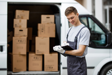 A young handsome smiling worker wearing uniform is standing next to the van full of boxes holding a clipboard in his hands. House move, mover service.の写真素材