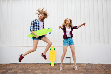 Two pretty blond girls wearing checkered shirts and denim shorts are jumping and dancing with bright longboards. Young girls are capering and having fun.の写真素材