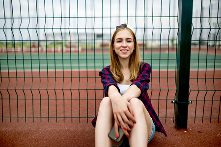 A pretty smiling blond girl wearing checkered shirt, white cap and denim shorts is sitting leaned on the guardlattice on the sports field with a phone in her hands. Sport and cool style.の写真素材