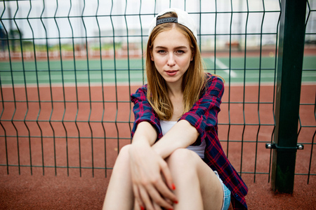A pretty smiling blond girl wearing checkered shirt, white cap and denim shorts is sitting leaned on the guardlattice on the sports field. Sport and cool style.の写真素材
