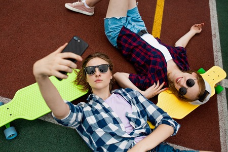Two pretty blond girls wearing checkered shirts, caps and denim shorts are lying on the bright longboards on the sportsfield with their sunglasses on and making selfie. Sport and cool style.の写真素材