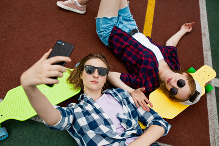 Two pretty blond girls wearing checkered shirts, caps and denim shorts are lying on the bright longboards on the sportsfield with their sunglasses on and making selfie. Sport and cool style.の写真素材