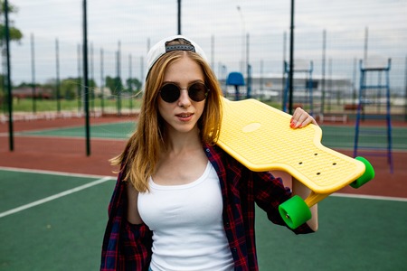 A pretty smiling blond girl wearing checkered shirt, white cap and sunglasses is standing in front of the guardlattice on the sports field with a yellow longboard on her shoulder. Sport and cool style.の写真素材