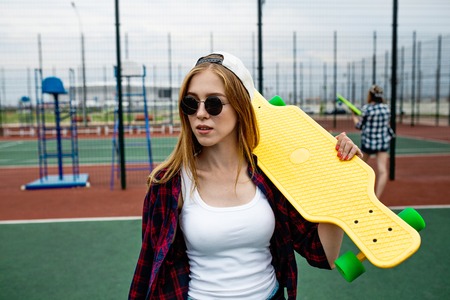 A pretty smiling blond girl wearing checkered shirt, white cap and sunglasses is standing in front of the guardlattice on the sports field with a yellow longboard on her shoulder. Sport and cool style.の写真素材