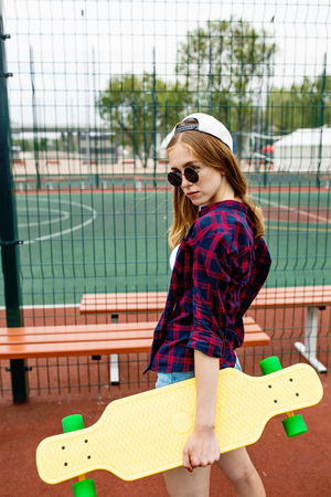 A pretty blond girl wearing checkered shirt, white cap and sunglasses is standing on the sports field with a yellow longboard in her hand. Sport and cool style.の写真素材