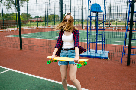 A pretty blond girl wearing checkered shirt, white cap and sunglasses is walking through the sports field with a yellow longboard in her hands. Sport and cool style.の写真素材