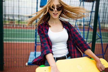 A pretty smiling blond girl wearing checkered shirt, white cap and sunglasses is walking through the sports field with a yellow longboard in her hands. Flying hair. Sport and cool style.の写真素材