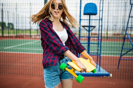 A pretty smiling blond girl wearing checkered shirt, white cap and sunglasses is walking through the sports field with a yellow longboard in her hands. Flying hair. Sport and cool style.の写真素材