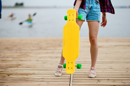 A girl wearing denim shorts is standing on the pier and holding a yellow longboard in front of her. River is in the background.の写真素材