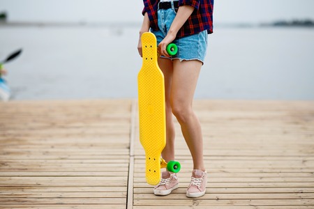 A girl wearing denim shorts is standing on the pier and holding a yellow longboard in front of her. River is in the background.の写真素材