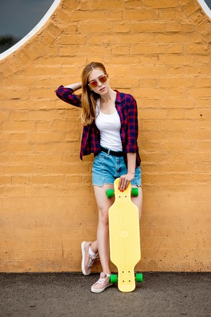 A girl wearing sunglasses, checkered shirt and denim shorts is standing in front of the brick wall with her hand behind her head and holding a yellow longboard in front of her. Cool style.の写真素材